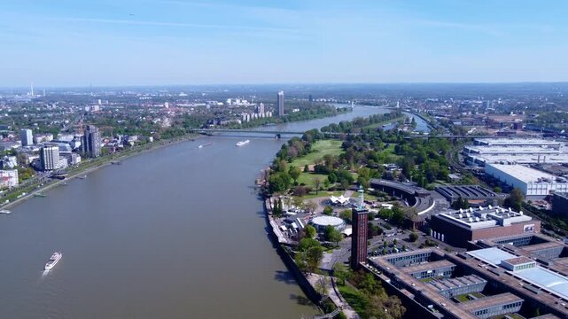 Drone aerial of Cologne Rhine riverfront with Rheinpark Tower, bridges, green parkland and boats. Clear daytime cityscape showing urban development and waterfront infrastructure.