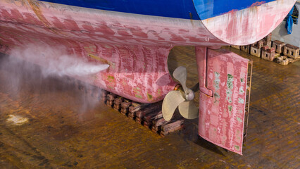 massive cargo ship hull, propeller, and rudder in a dry dock during maintenance. High-pressure water jet cleaning the red surface. Marine engineering, shipyard repair, heavy industry. © Yellow Boat