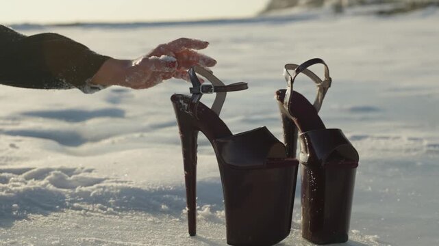 Frozen female hand in the snow reaches for pair of high-heeled women's dance shoes standing in the snow during the day.