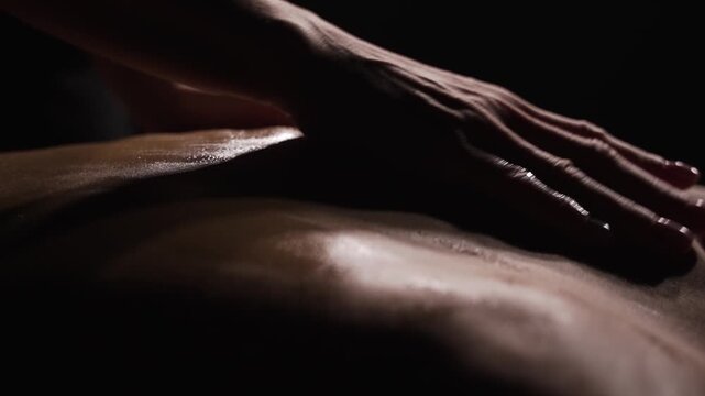Close-up of masseuse giving shoulder massage to a young, slender woman in the dark. The masseuse's hands are at work.