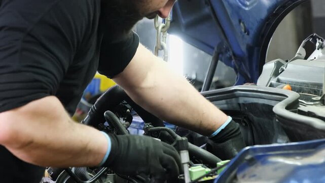 Mechanic performs engine repair tasks touching parts with hands. Specialist of car service rebuilds engine standing against chain hook
