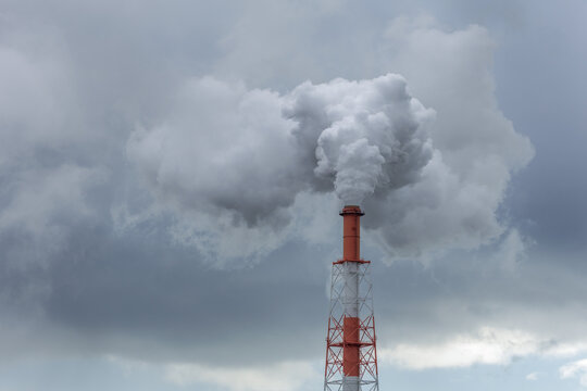 Tall red and white industrial chimney releases a dense, billowing plume of white-gray smoke into a heavy, grey overcast sky, illustrating air quality, industrial emissions, and environmental impact.