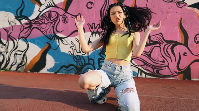 Trendy young female dancer listening to music and performing energetic street dance routine dynamic hip hop moves outdoors, against colorful graffiti wall, freedom and lifestyle, and creative energy