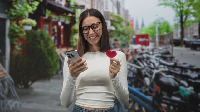 Young woman smiling while holding a smartphone and a small red heart, looking at her phone on a city street; romance dating connection.