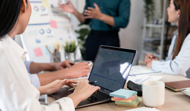 A business professional presents financial data and strategy on a laptop during a team brainstorming session in a modern office, featuring a whiteboard background.