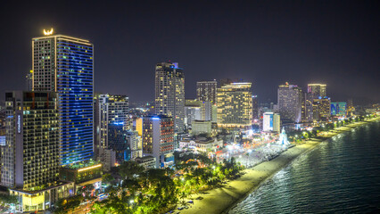 Naklejka premium Night view of Nha Trang, Vietnam showcasing a bustling beachfront illuminated by city lights. The skyline of modern high-rise buildings glows against the dark sky, while the lively promenade