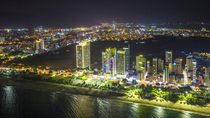 Obraz premium Night view of Nha Trang, Vietnam showcasing a bustling beachfront illuminated by city lights. The skyline of modern high-rise buildings glows against the dark sky, while the lively promenade