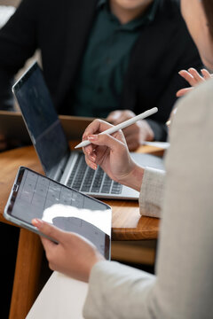 Professional woman using a stylus on a digital tablet, collaborating with a colleague on a laptop during a modern business meeting in an office. Vertical orientation.