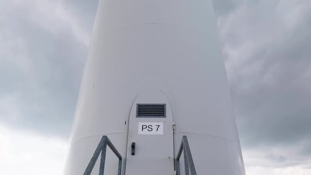 The base of a wind turbine labeled 'PS 7' stands against a cloudy sky, showcasing renewable energy infrastructure. Useful for environmental documentaries or educational content.