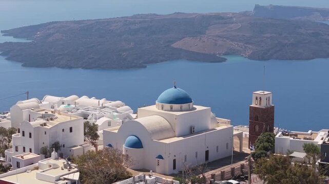 Drone orbiting a white church with blue dome in Imerovigli, Santorini, revealing the volcanic caldera and the deep blue Aegean Sea in the background.