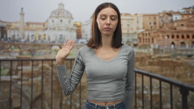Young hispanic woman raising hand with palm visible beside a metal railing in front of a historic building ruins, neutral expression; indifference.