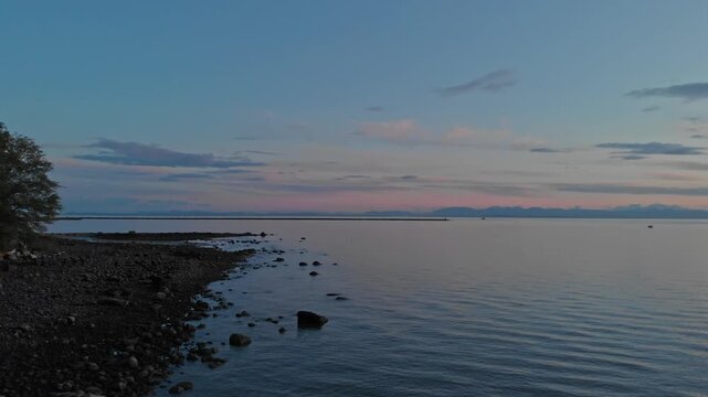 Smooth drone flight glides over calm sea surface tracking toward shoreline forest while vibrant pink sky reflects on water during twilight hours at University of British Columbia coastline
