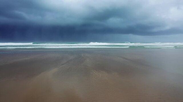 Slow motion static pan of ocean waves rolling and receding on sandy shoreline with dark storm clouds over coastal beach at overcast evening