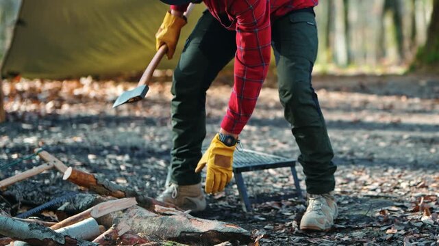 Camper man chopping firewood with a sharp axe on a wooden log in a forest campsite during daytime.