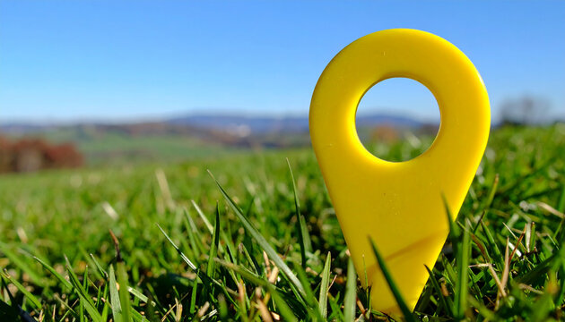 Vibrant yellow location pin standing upright in lush green grass blades, rolling hills and bright blue sky, travel and exploration concept