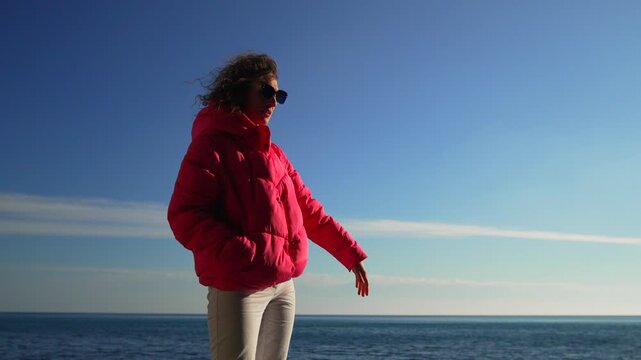 Beach pink jacket woman wearing a bright winter coat waves while looking at the sea under a clear blue sky on vacation