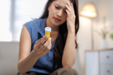 Fototapeta premium Asian woman feeling unwell with a migraine, holding a medicine bottle, experiencing pain and stress at home