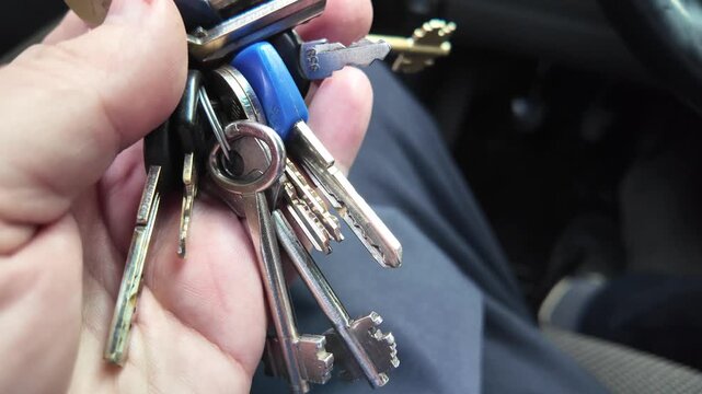 A male hand holding a large bunch of various metal keys on a keyring inside a car. Close-up view of house keys against a blurred vehicle interior background.