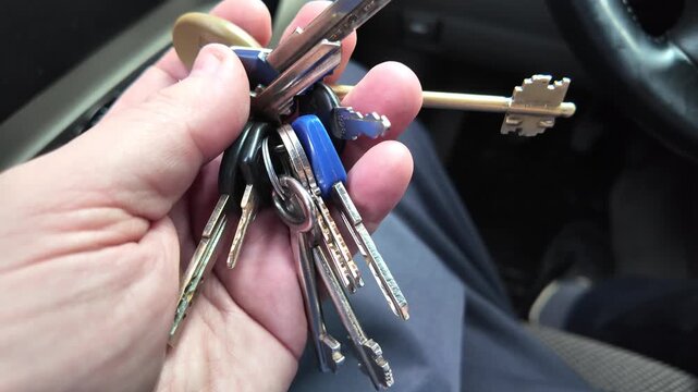 A male hand holding a large bunch of various metal keys on a keyring inside a car. Close-up view of house keys against a blurred vehicle interior background.