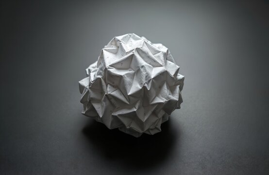 White crumpled paper ball with small symbols printed on surface rests on dark gray surface. Paper texture shows many folds, creases. Object appears abstract, textured, suggesting creative blockage
