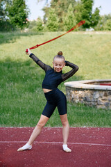 Fototapeta premium Young girl performs rhythmic gymnastics with a ribbon on a sports field outdoors, showcasing athletic skills. Young Girl Practicing Rhythmic Gymnastics Outdoors on Sunny Day