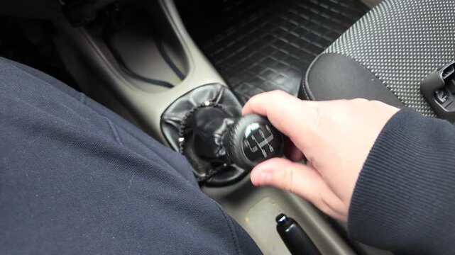 Close-up of a driver's hand shifting gears on a black leather manual transmission stick inside a car. A male hand holds the gearstick, controlling the vehicle. Automotive interior details.