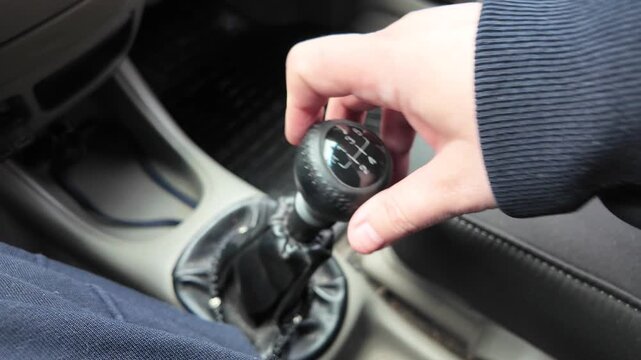 Close-up of a driver's hand shifting gears on a black leather manual transmission stick inside a car. A male hand holds the gearstick, controlling the vehicle. Automotive interior details.