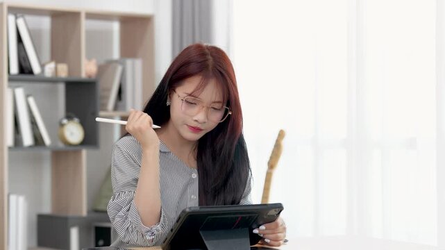 Young woman with long hair using digital tablet to create art or design while sitting at modern minimalist workspace with wooden table in bright room.