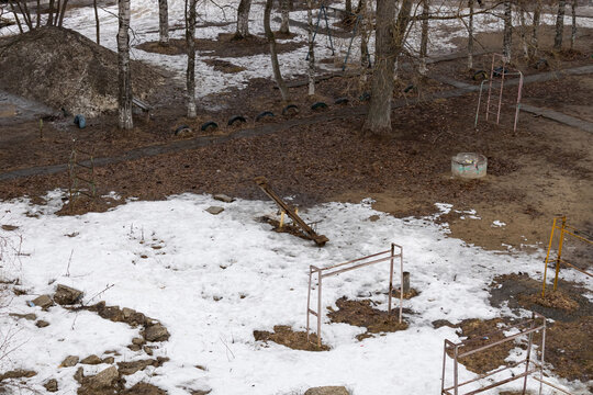 High-angle view of a worn playground with a seesaw in a Russian northern residential area. Patches of melting snow, dark muddy earth, and bare birch trees mark the beginning of spring. 