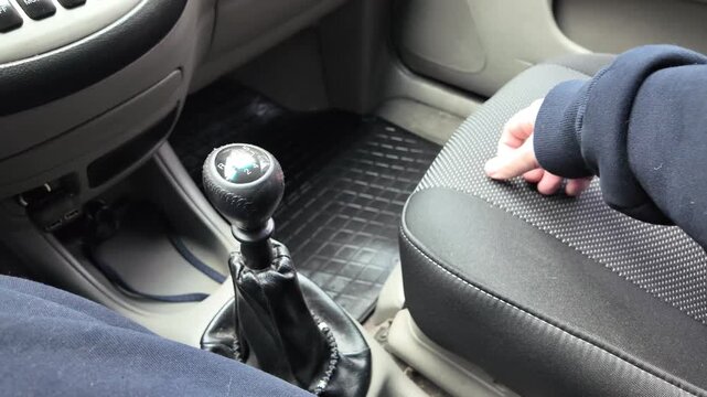 Close-up of a driver's hand shifting gears on a black leather manual transmission stick inside a car. A male hand holds the gearstick, controlling the vehicle. Automotive interior details.
