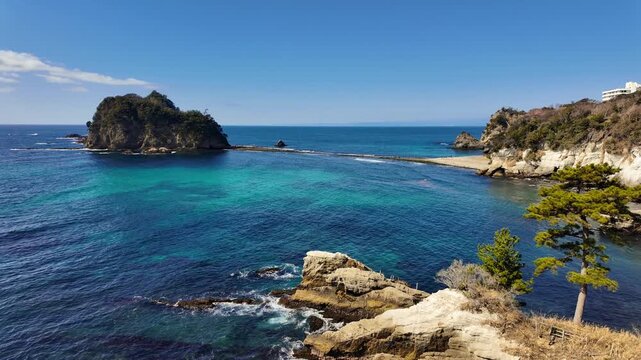 A high-angle panoramic tilt and pan across the crystal-clear turquoise waters of Dogashima, Japan. The shot captures the rugged volcanic cliffs, a lone pine tree