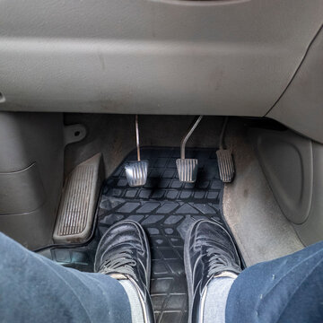 Close-up of driver's feet in black shoes resting on a rubber floor mat near metal pedals inside a manual transmission car. Automotive interior detail showing clutch, brake and gas control.