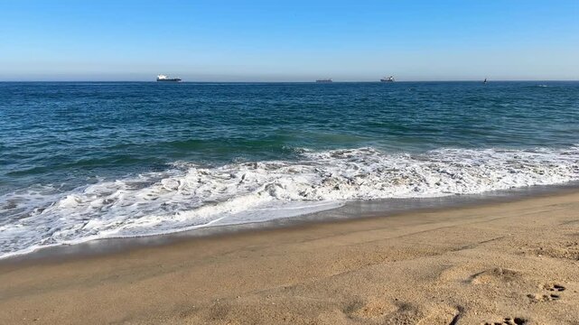 Powerful sea waves hitting the beach with white foam, featuring three container ships on the distant horizon and wet sand reflections. Industrial and nature elements in one shot.