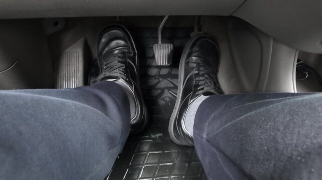 Close-up of driver's feet in black shoes resting on a rubber floor mat near metal pedals inside a manual transmission car. Automotive interior detail showing clutch, brake and gas control.