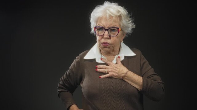 Elderly woman with white hair and colorful glasses, hand on chest and pursed lips, red nails and ring, brown sweater with white collar in studio; concern vulnerability.
