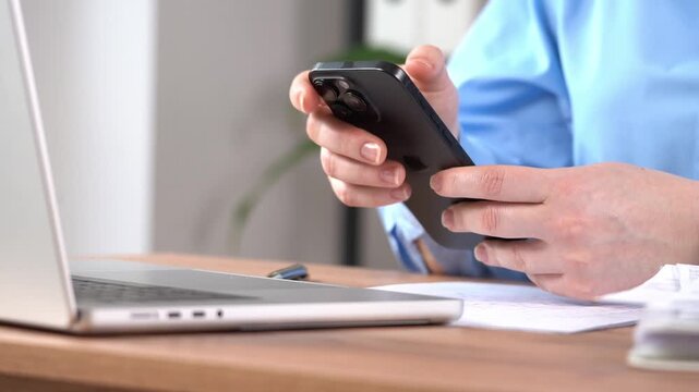 Hands hold smartphone. Fingers scroll on touchscreen. Laptop open on wooden desk. Papers lie nearby. Person wears blue shirt. Calm and focused expression