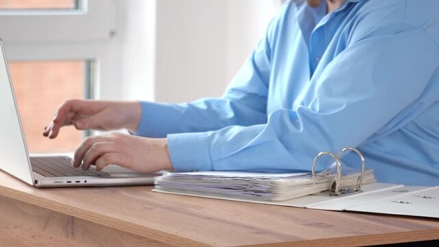 Close-up of hands typing on laptop. Fingers move quickly across keys. Person in blue shirt sits at wooden desk. Binder and papers lie open. Desk features binder, documents, and laptop