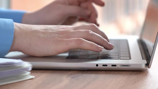 Close-up of hands typing on a laptop keyboard. Person's arm in blue shirt, focused on work. Wooden desk with stacked papers, blurred office background