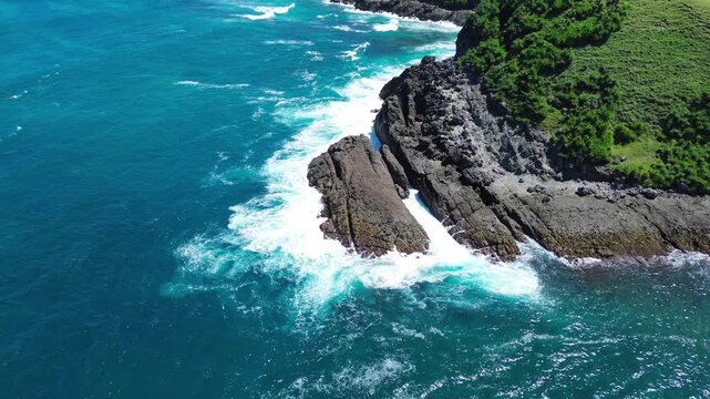 An aerial shot shows waves crashing along a rugged coastline in Gunungkidul Regency, Yogyakarta, Indonesia. Lush greenery covers the rocky cliffs meet