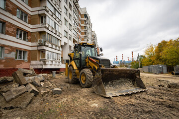 Fototapeta premium A backhoe is digging and moving earth near a residential building. Workers are focused on the job site as the sky is cloudy. Fences and debris are visible