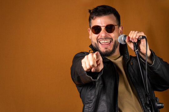 Hombre rockero con chaqueta de cuero y lentes cantando en&eacute;rgicamente con su microfono y se&ntilde;alando a c&aacute;mara