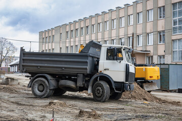 Obraz premium A dump truck is parked on a construction site surrounded by machinery, dirt piles, and a large building under overcast skies. Workers are present in the area