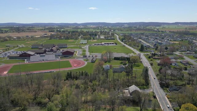 High angle drone footage showing a paved road winding past a red running track and athletic field surrounded by trees.