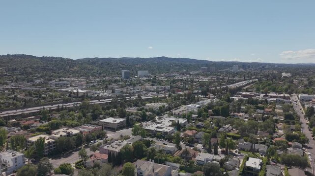 Aerial view of Sherman Oaks, Los Angeles with busy highway traffic cutting through residential neighborhoods, surrounded by green trees and hills under clear sunny California sky. Panorama.