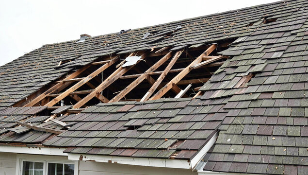 Damaged roof shows exposed wooden beams and missing shingles. Rooftop has large hole with broken structure visible. House siding and window frame are partially shown below