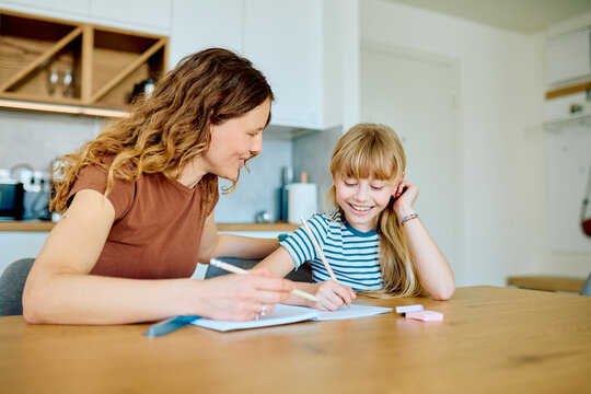 Mother and daughter are smiling while doing school homework with pencils and ruler at a wooden table in their bright kitchen