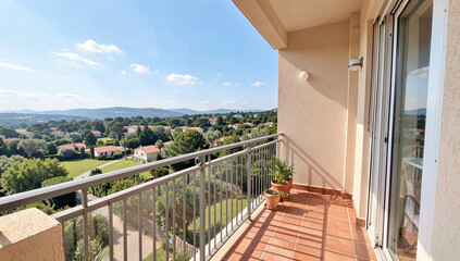 Obraz premium Balcony overlooks green hills and distant buildings. Bright blue sky with scattered clouds above. Metal railings frame the tiled balcony area. Potted plant sits near glass doors on the balcony