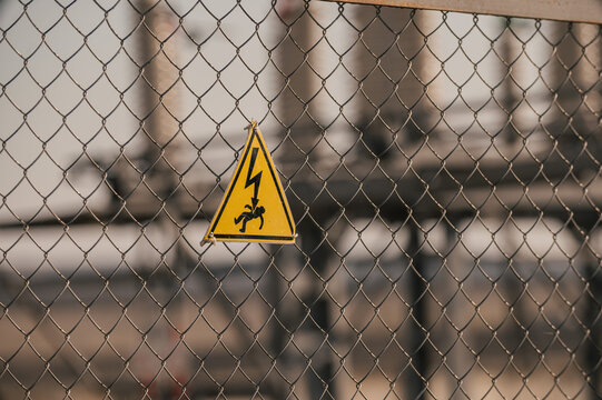 High voltage warning sign attached to metal fence in front of electrical substation. Blurred industrial background with power equipment, safety symbol, restricted area and electricity hazard concept.