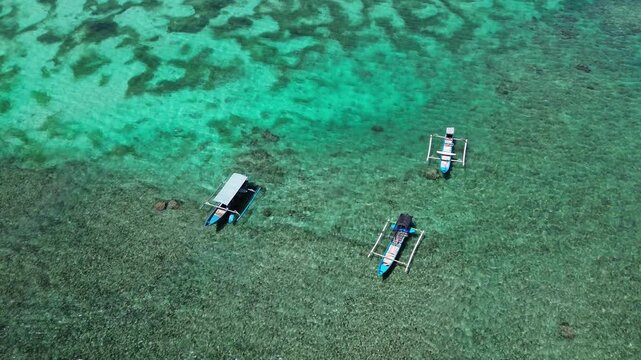 An aerial view shows three small outrigger boats floating in the shallow, clear, turquoise waters off the coast of a tropical island.