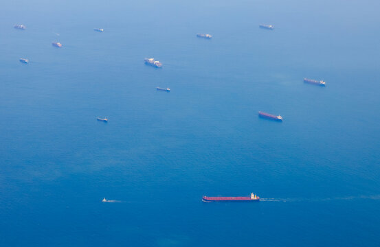 Multiple ships are seen on blue water. Some are far in the distance while others navigate closer to the viewer. Daylight creates a clear view of the scene. Ships wait to cross a strait.